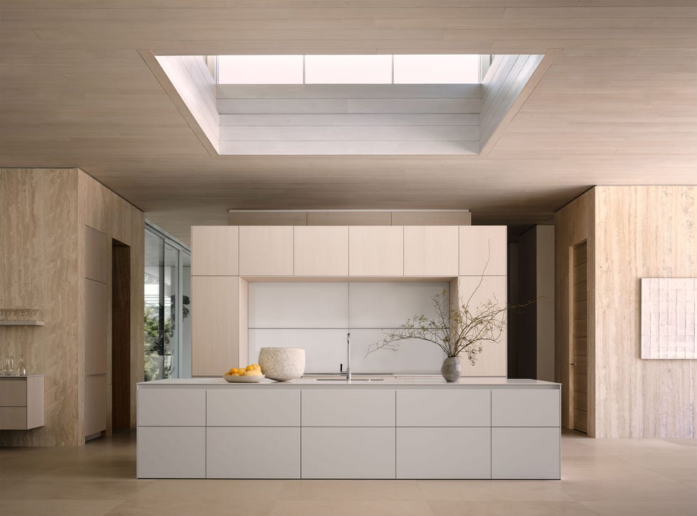Nicole Hollis - Elle Decor A light monochrome kitchen has a wall with cupboards and an island with a sink and a counter with vases and a bowl with oranges, corridors on both sides of the island, a large, square skylight and limestone floors
