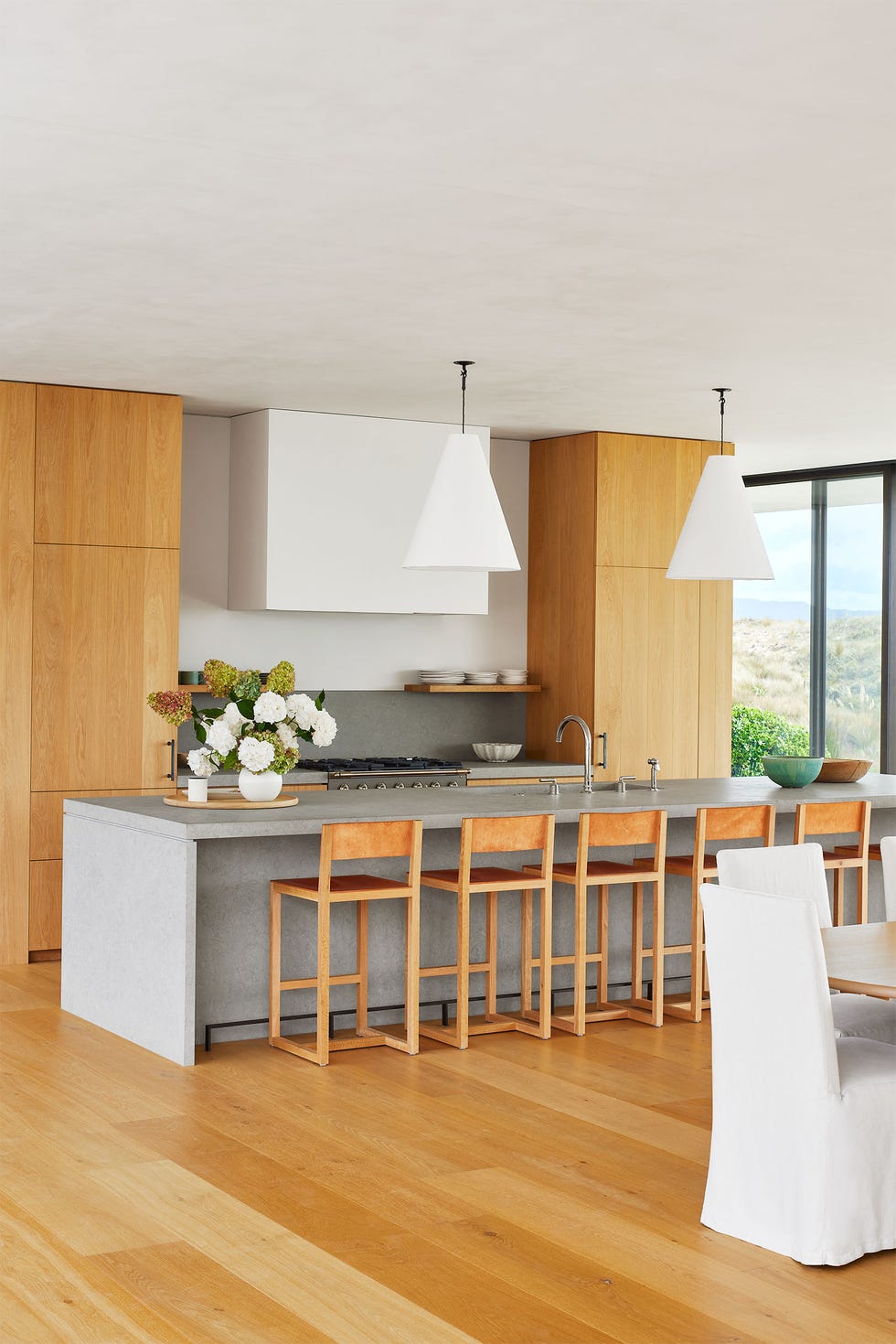 Jenny Kane - Elle Decor A kitchen with wooden floor and wooden floor to ceiling cabinets, gray limestone island with wooden hops, two large conical white pendants, glass wall