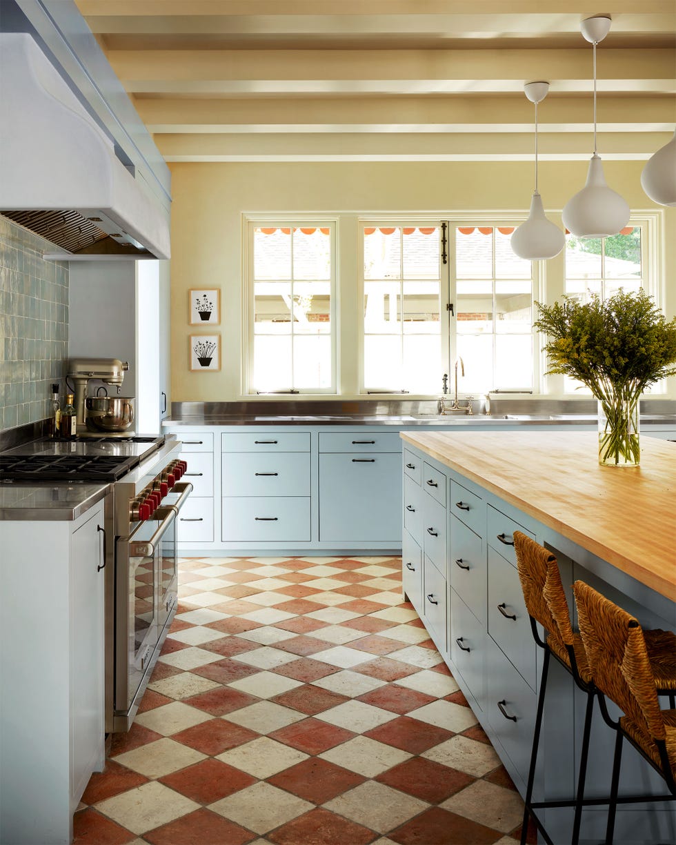 Ashe Leandro - Elle Decor Kitchen with light blue cupboards, bright yellow walls, island with butcher top and two rattan bridges and three tear pendants, windows above sinks, terracotta and white diamond shape tiles