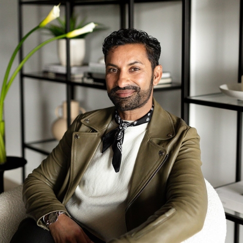 Picture of a man in a white shirt and a green leather jacket that sits in a white boucle chair. There is a black bookshelf behind him.
