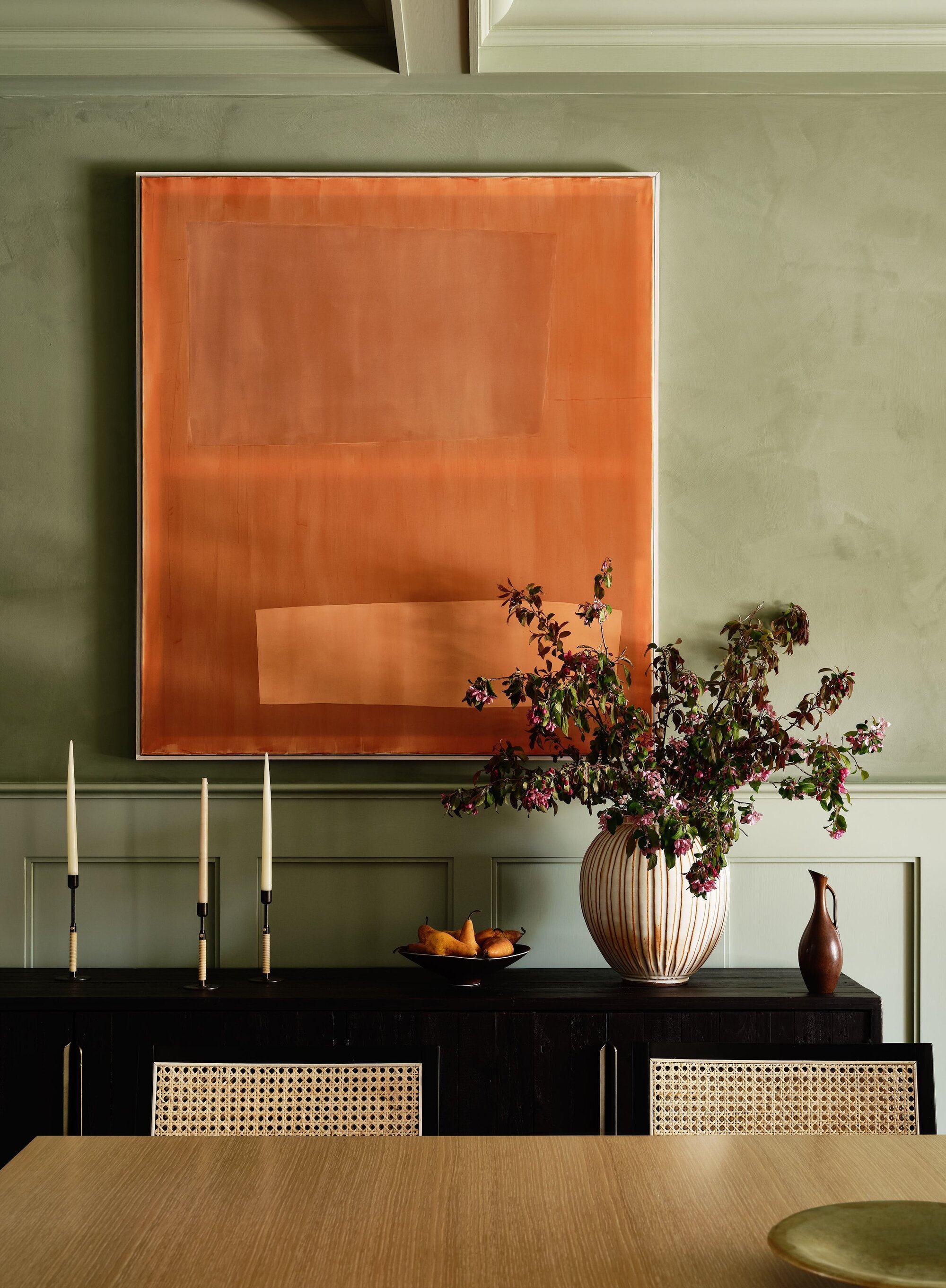 Image of a dining room that is designed by a wise green. There is a large orange painting on the wall with a black console table below. On the table there is a vase with purple flowers and three candle holders with candles.