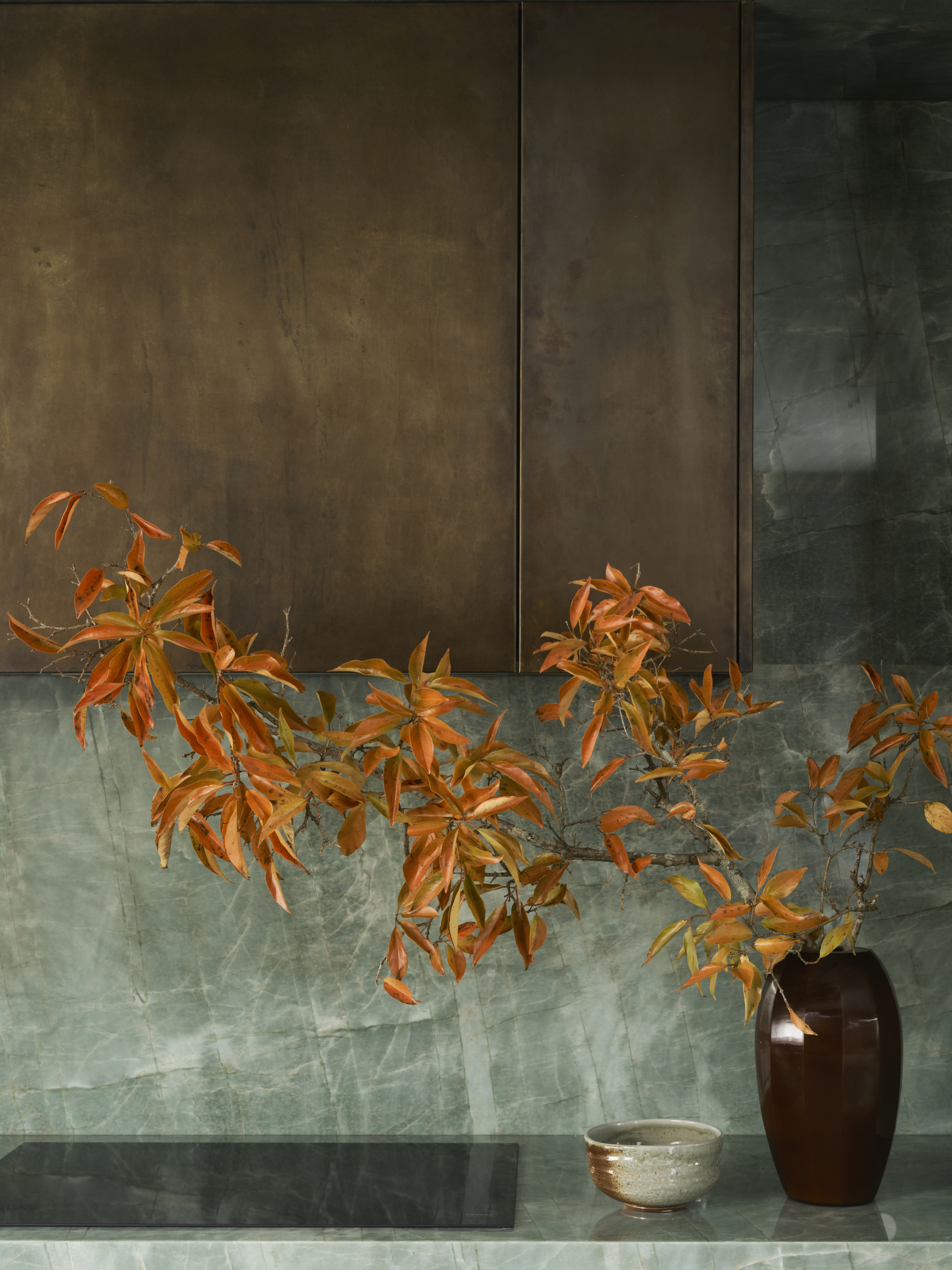 Close -up image of a kitchen counter and stove top with aged brass cabinets over it. The worktop and the back splash consist of a blue -green stone material. There is a vase made of bright orange flowers.