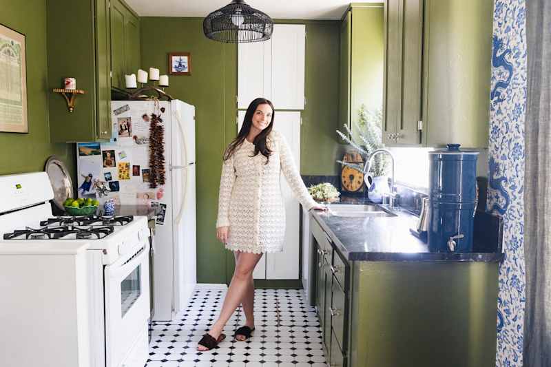A woman in a white lace dress is in a green kitchen with black worktops, a white stove and patterned curtains.