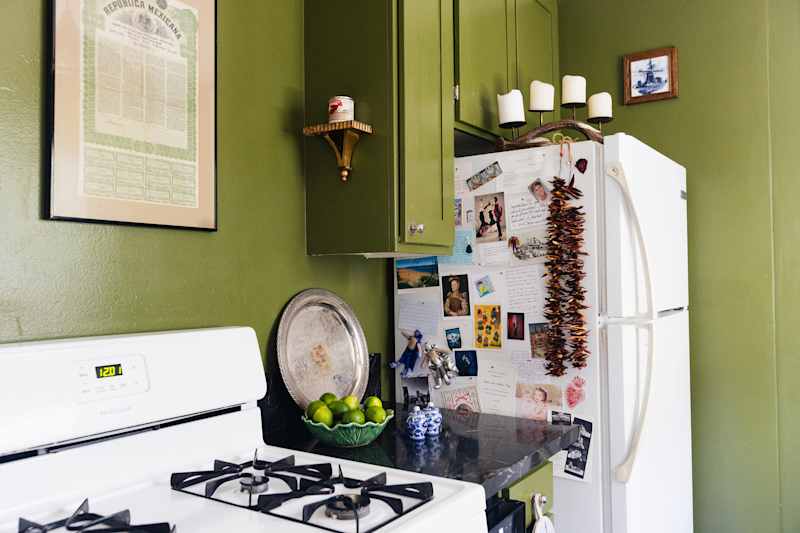 Kitchen corner with green walls, white stove, silver bowl, lime peel and a fridge covered with notes and photos.