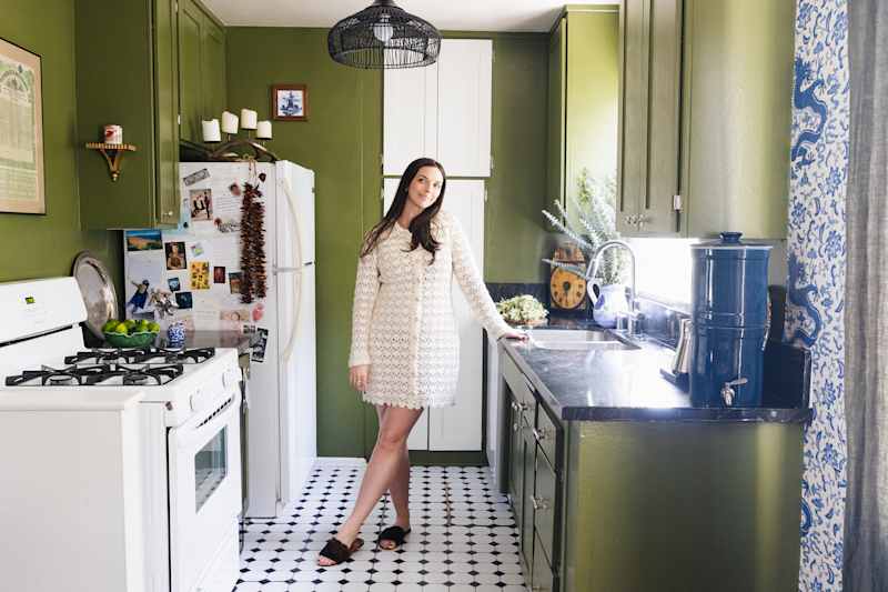 A woman in a white dress is in a green kitchen with black worktops, a gas stove and decorative objects.