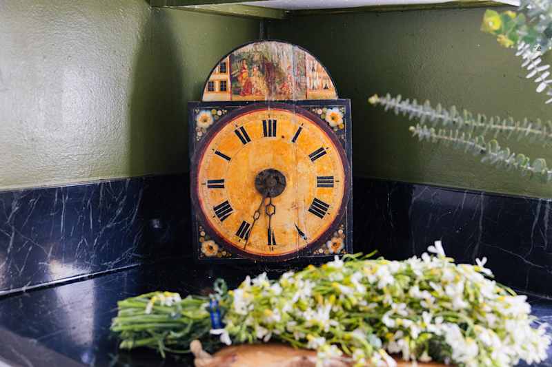 Vintage clock with yellow face and black Roman digits, surrounded by green and a dark marble surface.