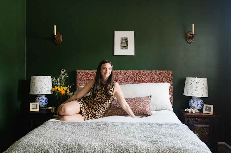 A woman in a leopard pressure dress sits on a bed with a green wall, flowery headboard and decorative lamps.