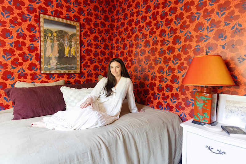 A woman in a white dress sits on a bed with beige bed linen, surrounded by living red flowers and a colorful lamp.