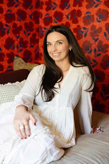 A woman with a long dark hair smiles while sitting on a couch and wearing a white dress against a red flowers.