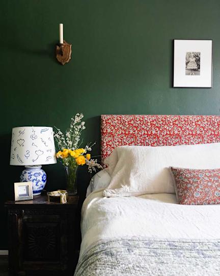 Cozy bedroom with a green wall, a red floral headboard, a white bed linen, a blue and white lamp and a vase made of yellow flowers.