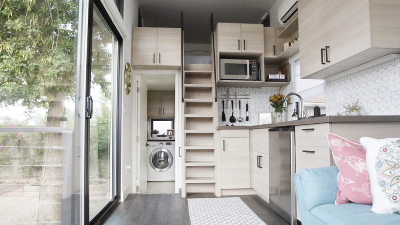 The inside of a tiny house with large glass windows, the kitchen, part of the living room and the loft bedroom.