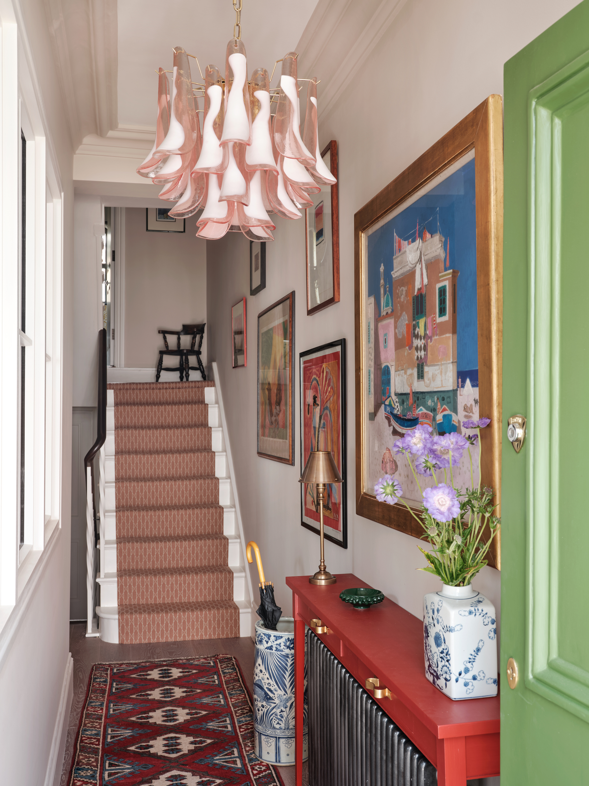 Home entrance with the gallery wall of the works of art, a red console table above a cooler and carpet with a pattern, a red patterned staircase and a sculptural pink glass trailer light