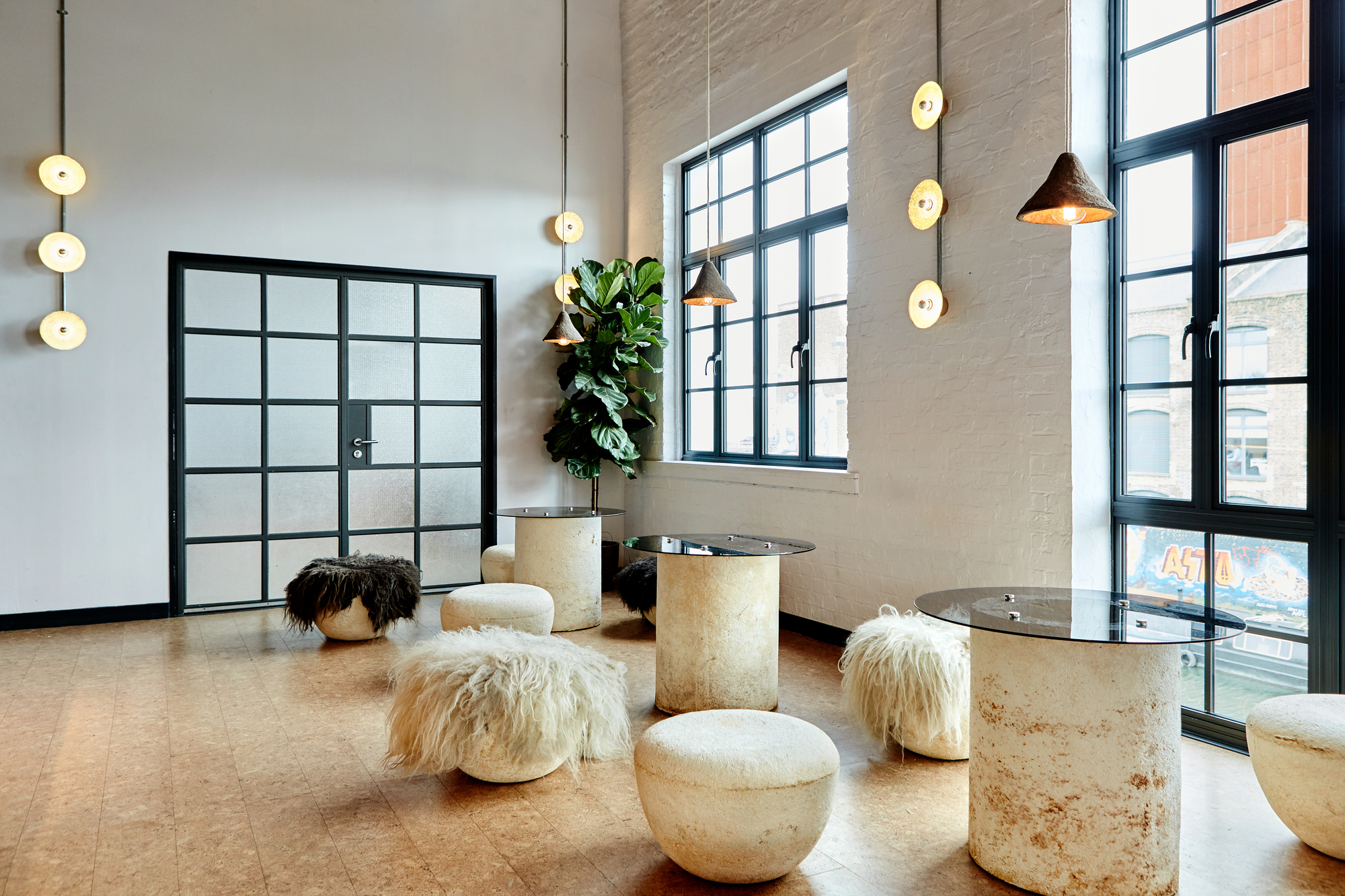 A cocktail bar with traverti tables with glass Upper table, cork floors, stone stools with sheepskin, large windows with steel frames, trailer lights and a pot fiddleaf in the corner