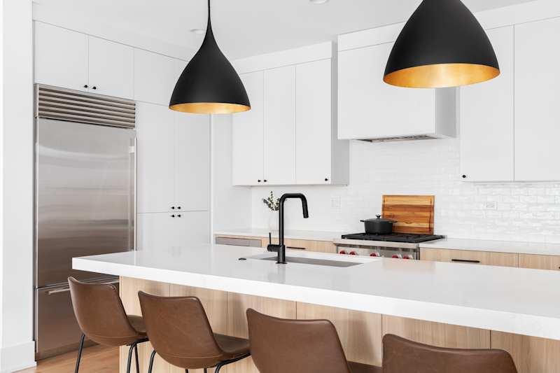 A kitchen detail with white and white oak cabinets, leather chairs on an island and a black and golden light that hangs above. No names or brands.