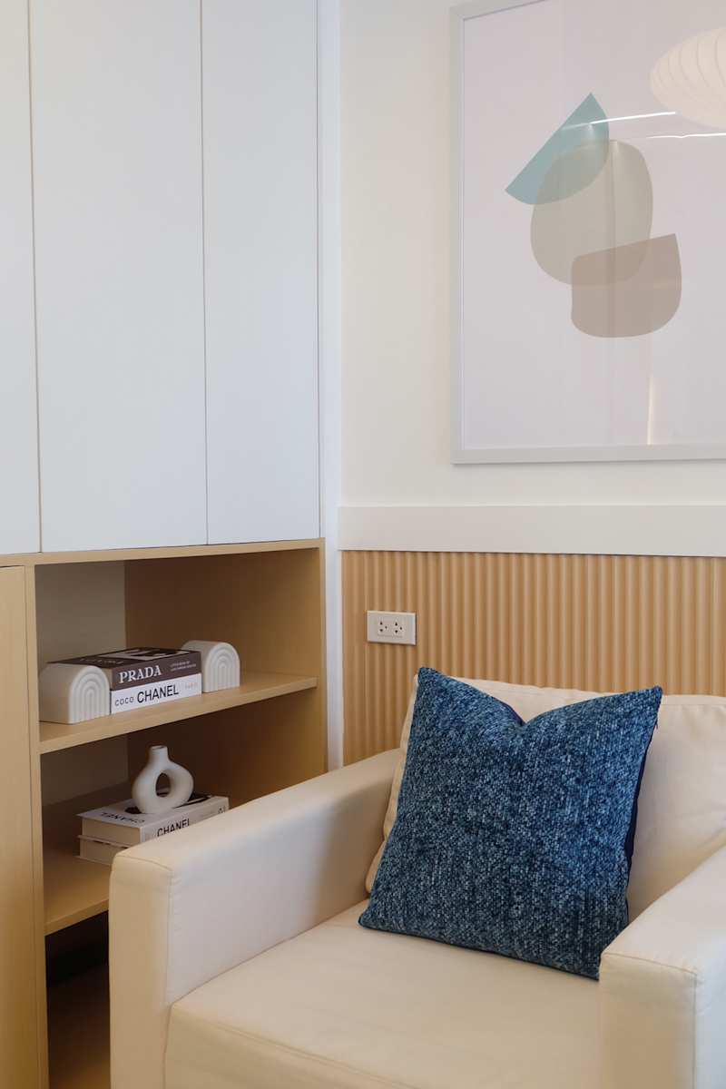 Cozy seating area with a white armchair with a blue structured pillow, a wooden shelf with books and abstract wall art.