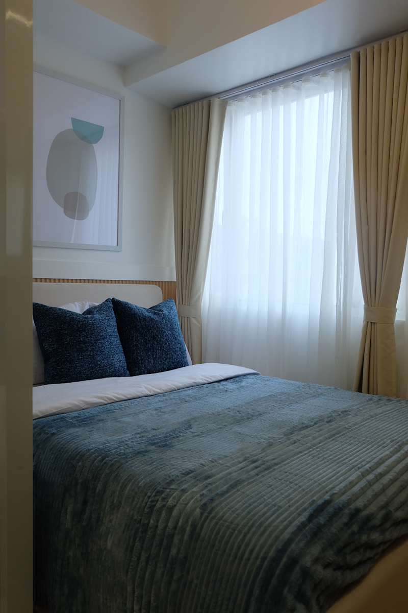 Cozy bedroom with a bed with a blue structured bed linen and decorative pillows, a large window with sheer curtains and abstract wall art.