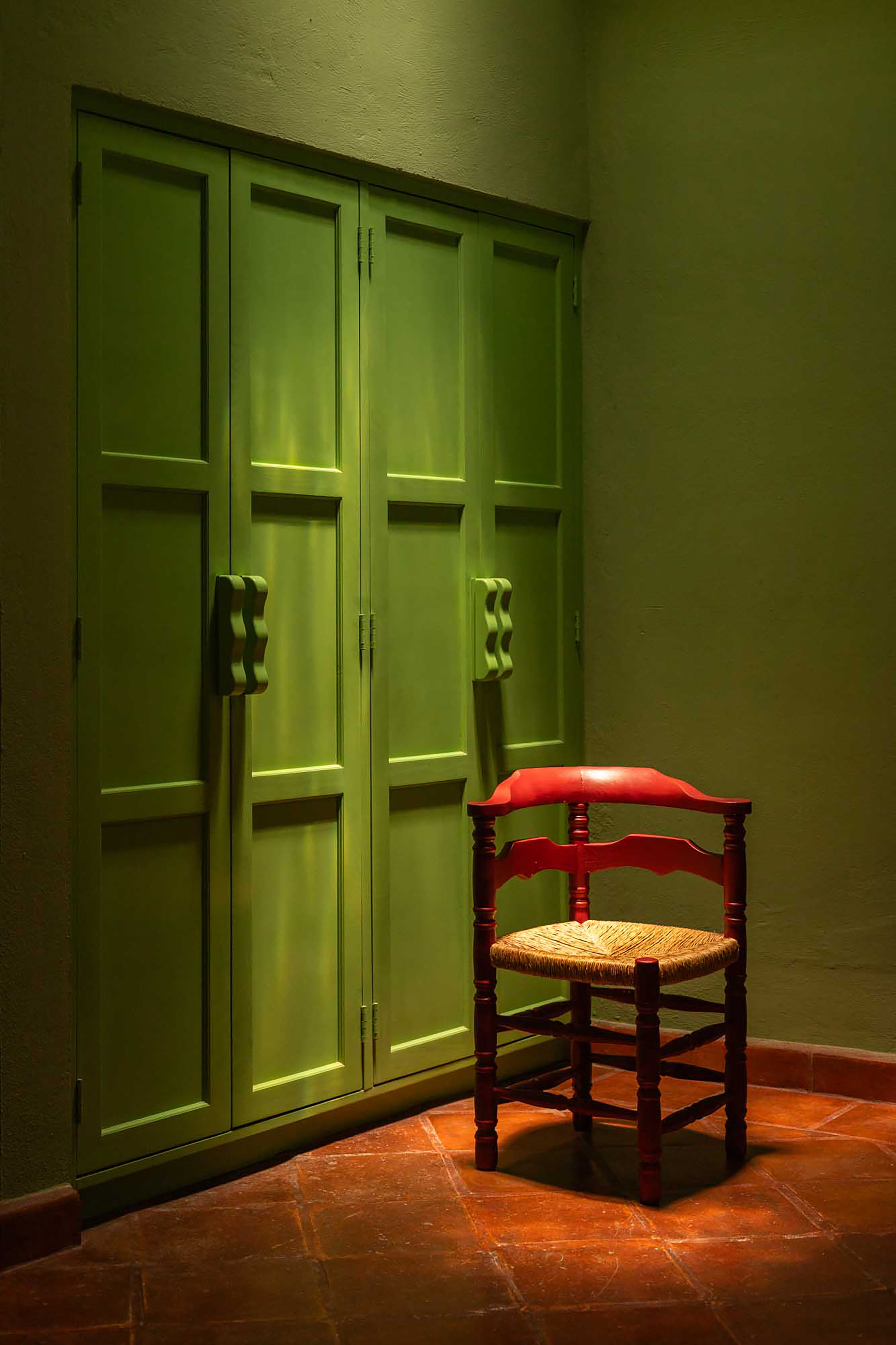 Corner of a room with orange terracotta floors and bright, olive green walls. There is a red chair in the shot.