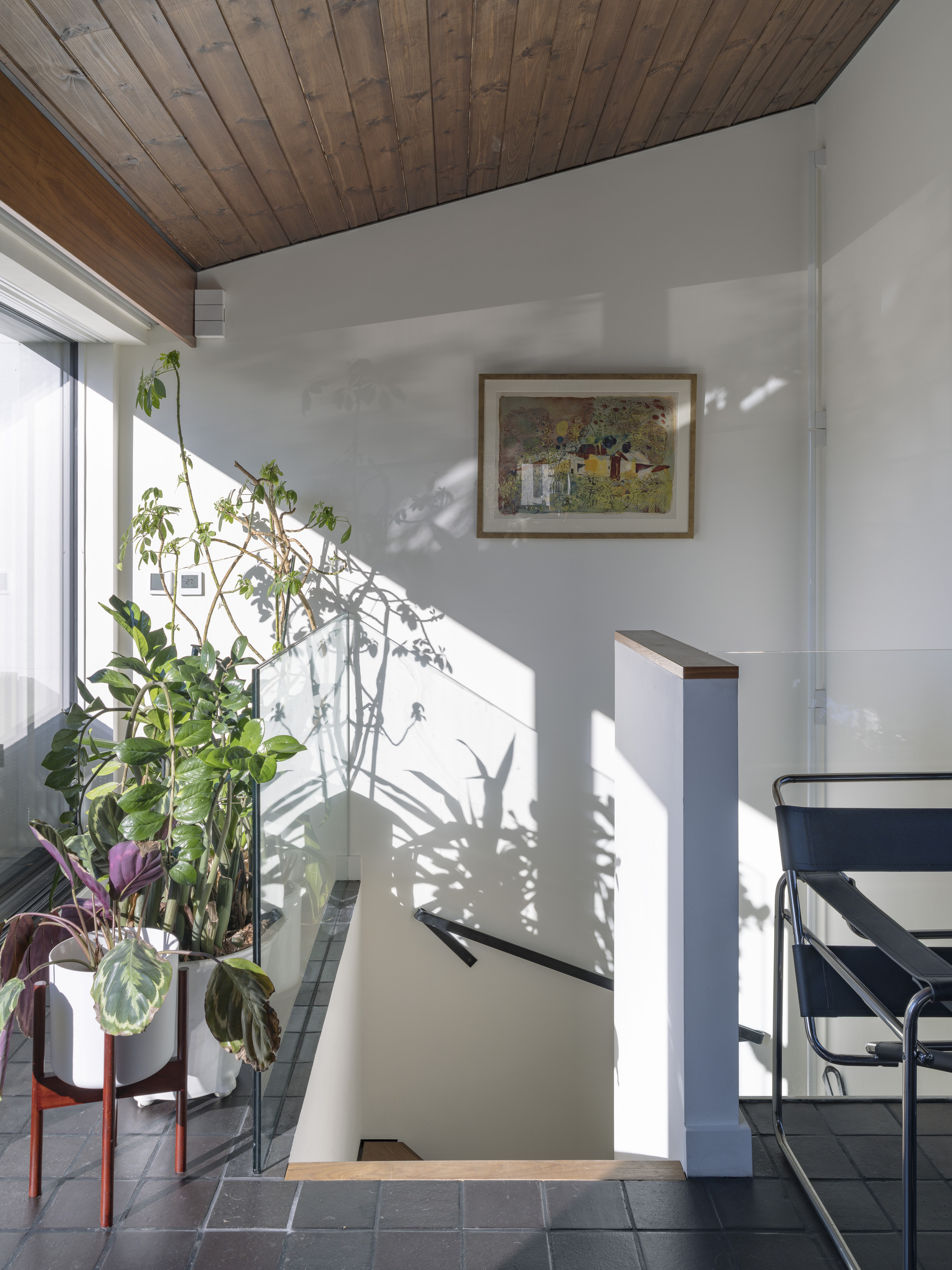 The top of a staircase in a house with a wooden ceiling, white walls, tiled floor, many plants in pots that sound in the sunlight, come through the window, a work of art that hung on the wall