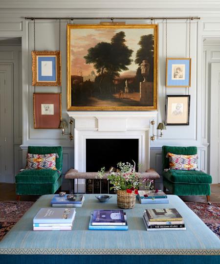 Living room with a large vintage landscape above the fireplace and smaller pictures on walls with two green velvet chairs and a large Ottoman Ottoman with books