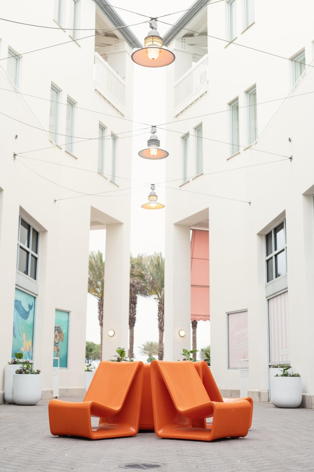 From Yeti to Seaside, this TX -based entrepreneur makes waves - 4 Four modern orange-colored chairs of tupelo goods are arranged in the middle of a sidewalk between two white buildings with large windows, skylights and palm trees in the background.