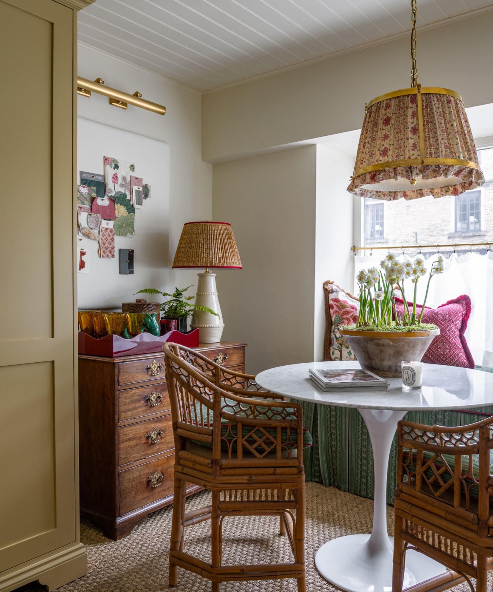 A corner of a room with a chest chest, above is a pretty lamp and decorative objects. There is a white gloss circular table with two bamboo -style chairs. On the table there is a diptyque candle and a rustic bowl with daffodils planted in moss. Sisal carpet on the floor. Neutral walls.