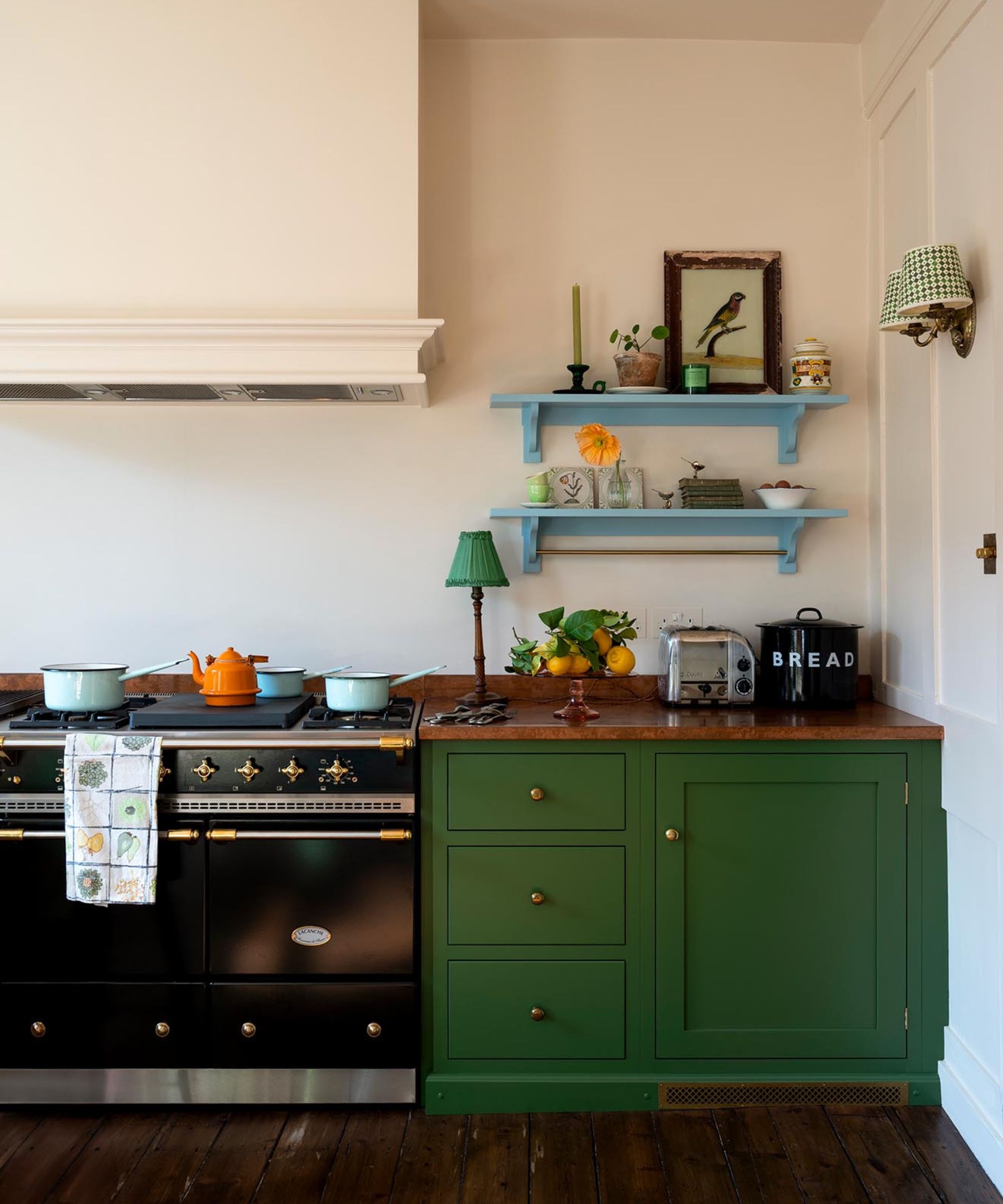 A small green kitchen with copper worktops and two blue floating shelves filled with vintage decor