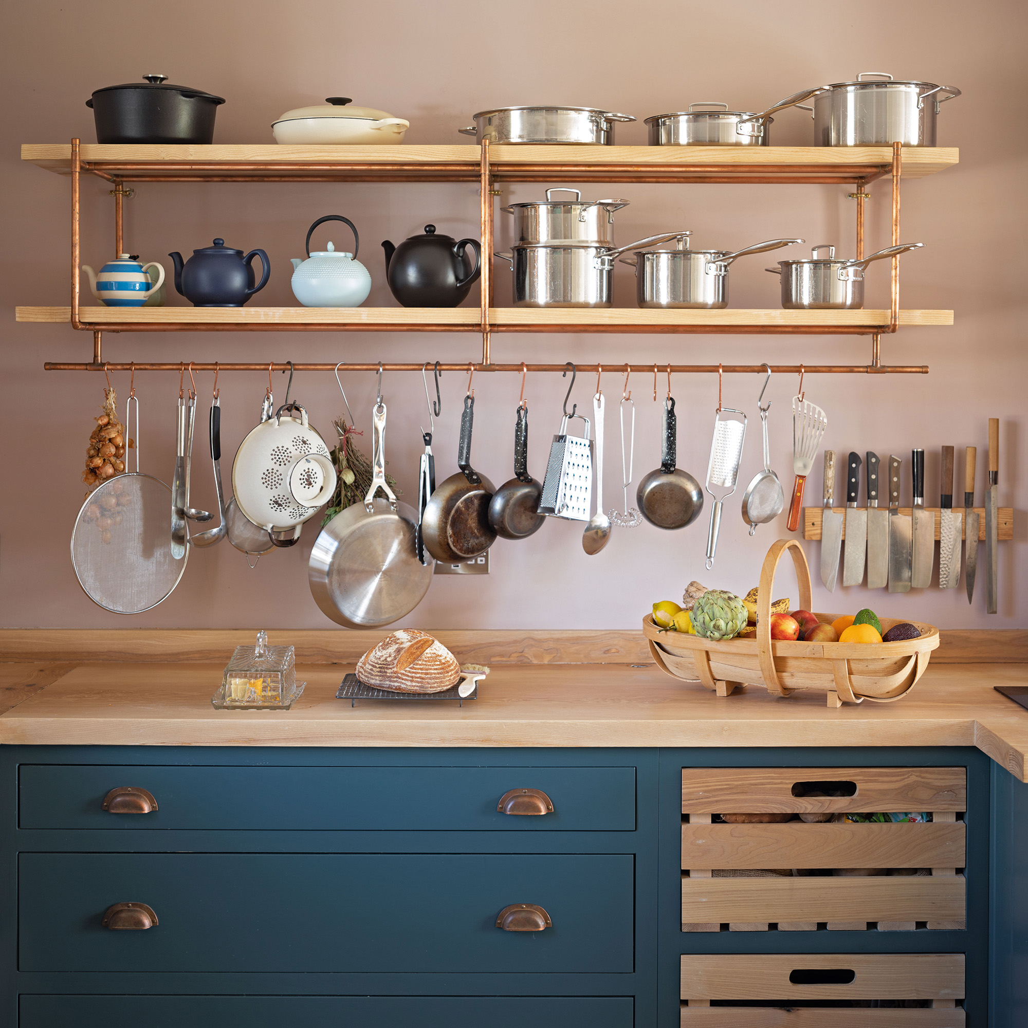 Country kitchen with pink wall and blue cupboards as well as copper and ash open shelves and hanging kitchen tools