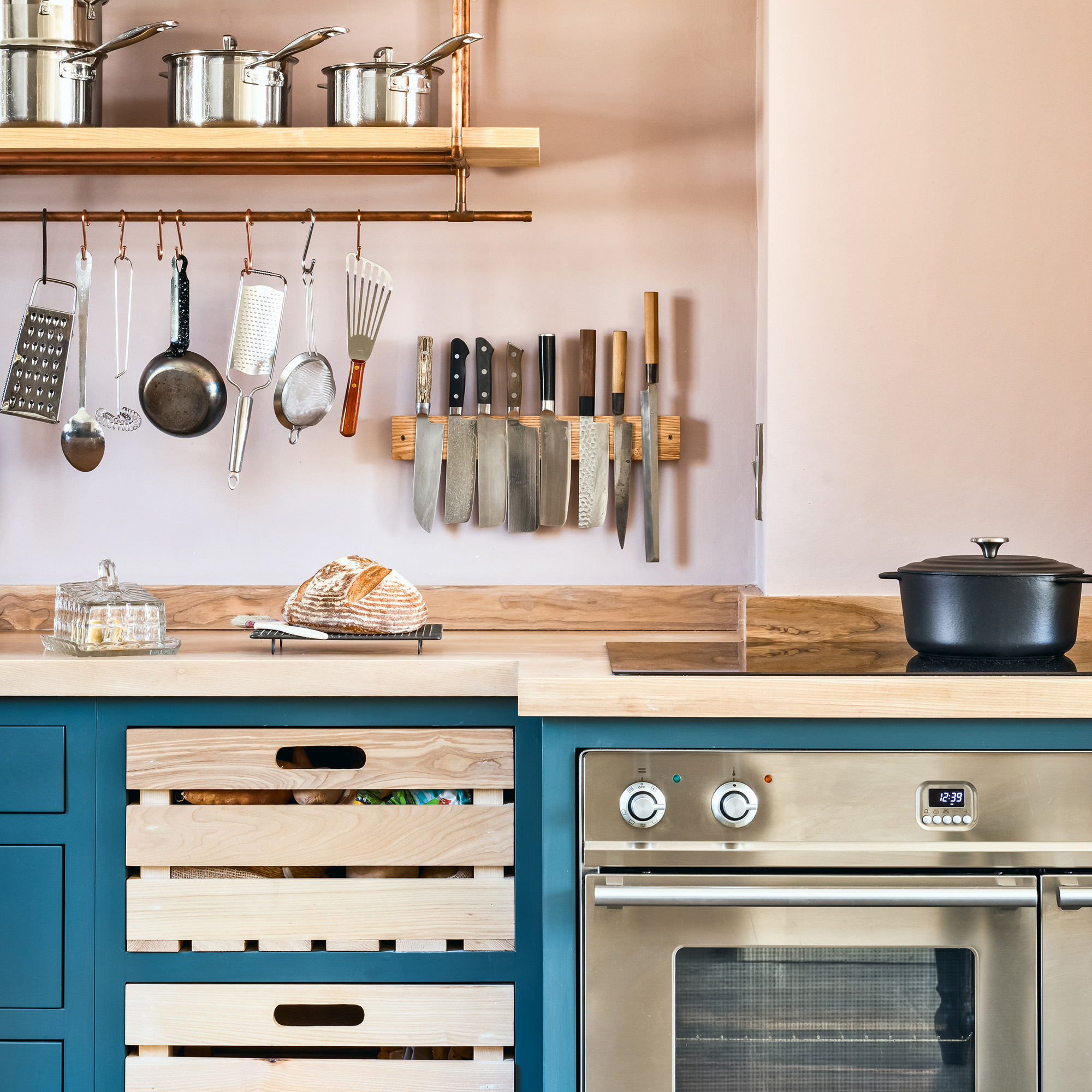 Country kitchen with pink walls and blue cupboards and with an open shelf made of copper pipe and ash, knife block and open boxes
