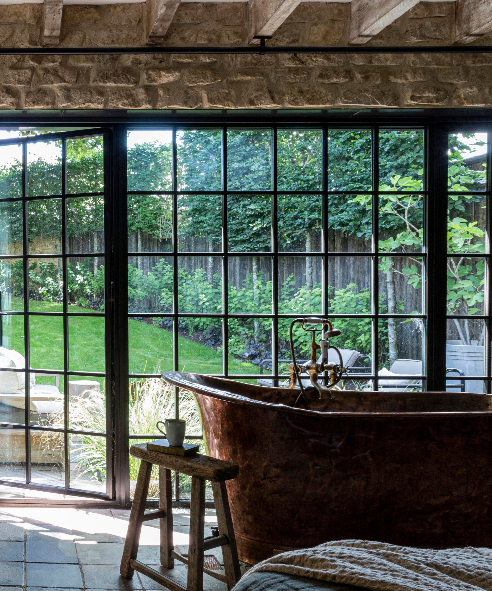 A bedroom with a copper bathtub in front of the window, styles with a wooden stool with a cup and a book crowned
