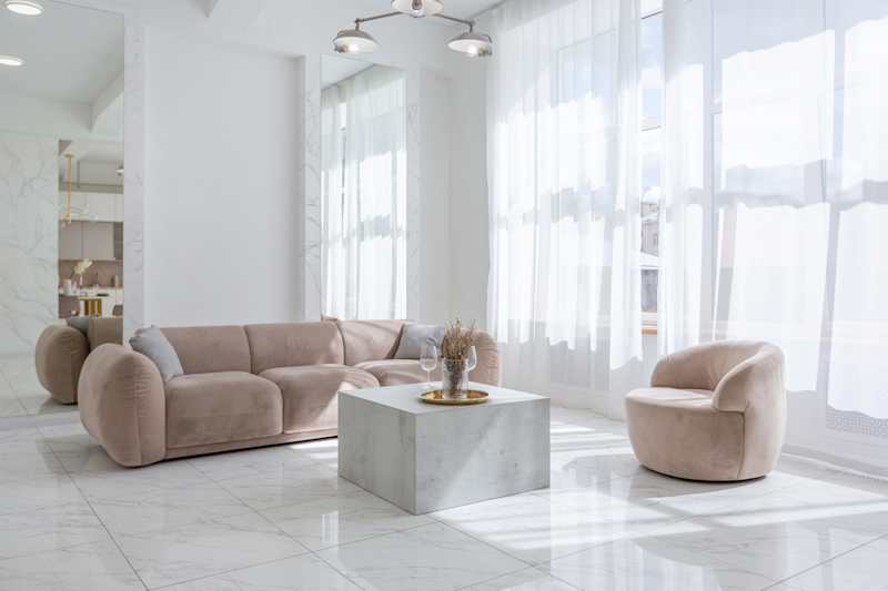 Living room with soft furniture in a light luxurious interior design of a modern apartment in a minimalist style with marble equipment and huge windows. Daylight inside.