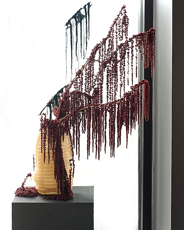 Vase with cascading red pearls on branches that are displayed against a window with dark stripes in the background.