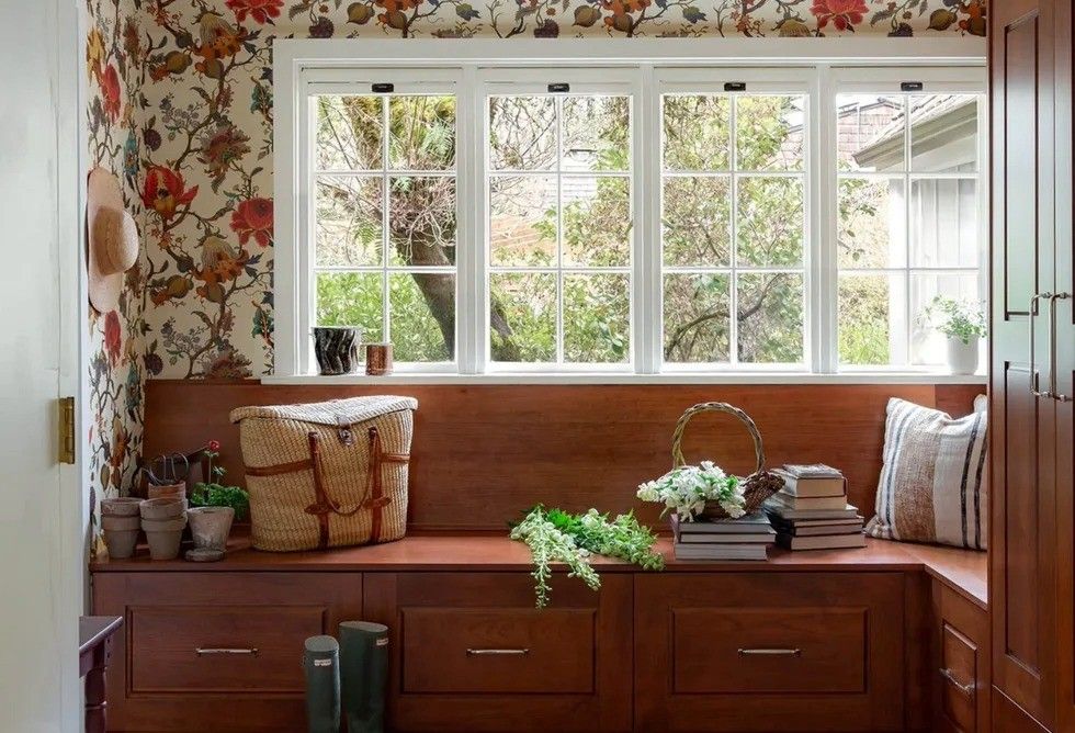 Cozy corner with floral wallpaper, window, bank, plants, books and a woven basket.
