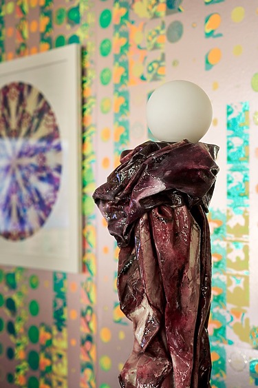 Abstract sculpture with a round top in front of colorful, patterned art on a wall.