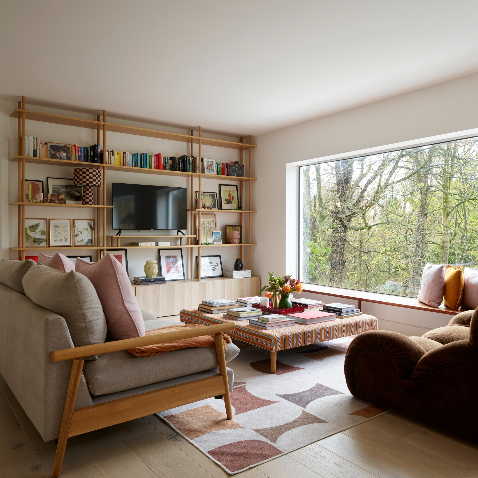 A living room with a large window, a gray sofa with a wooden frame, a brown sofa in the sofa style and an Ottoman coffee table