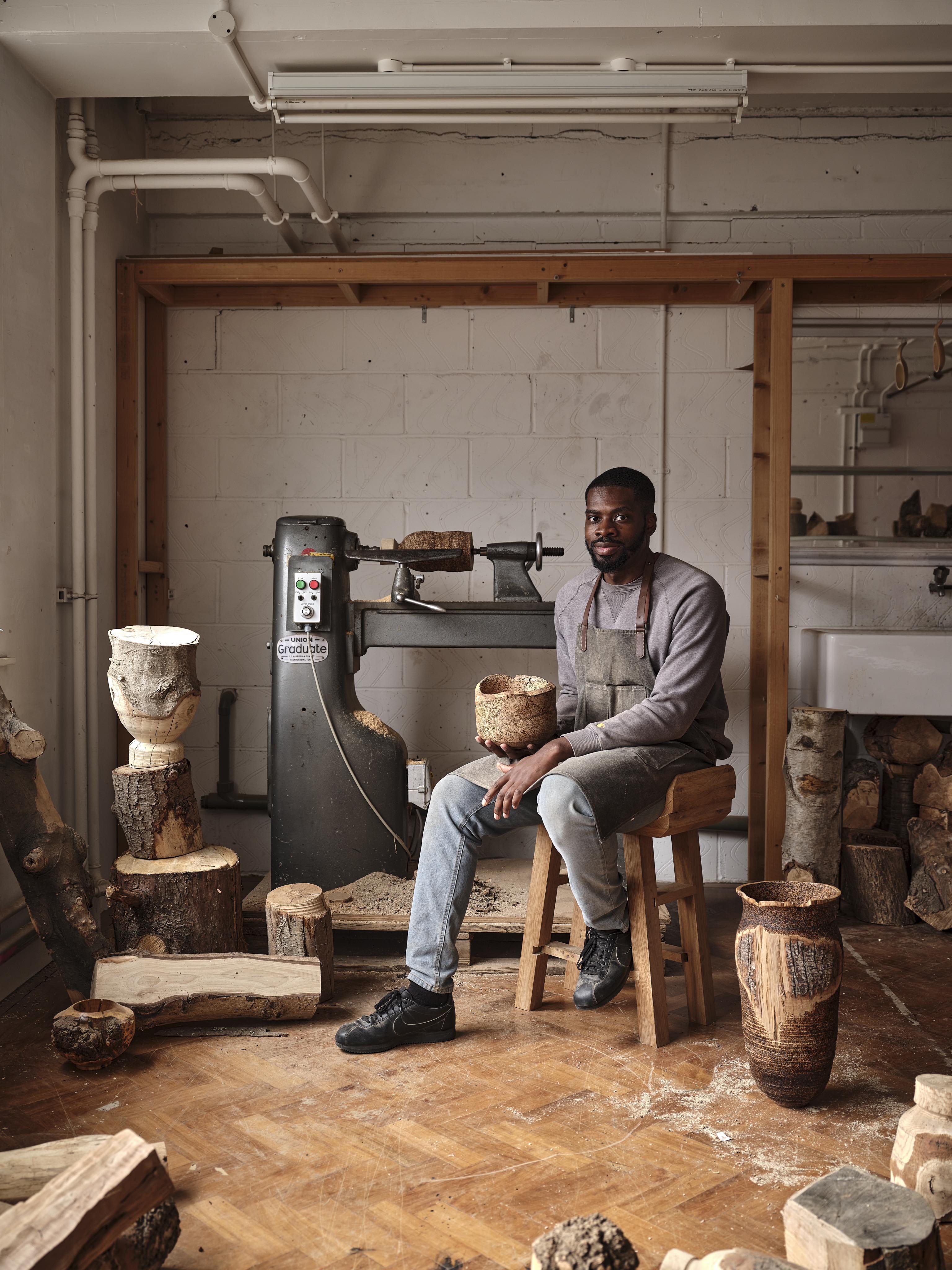 Artists, designers and craftsmen Darren Appiagiagei sit on a stool in a workshop next to the lathe, surrounded by wood sculptures