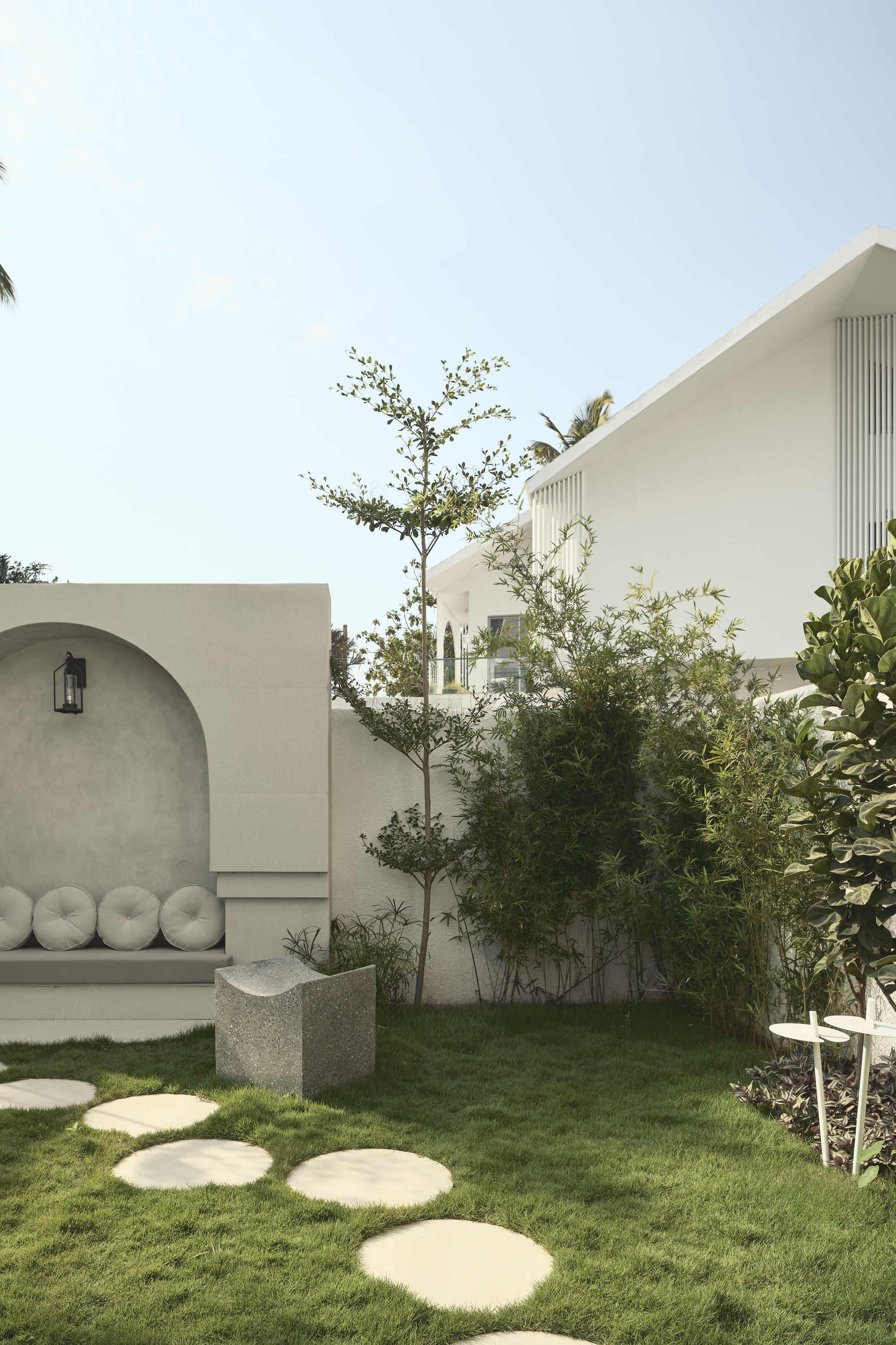 Image of a garden grasp with jumping stones that lead to a modern gray-be-bench seating area in an outer wall. There are also small trees along the wall.