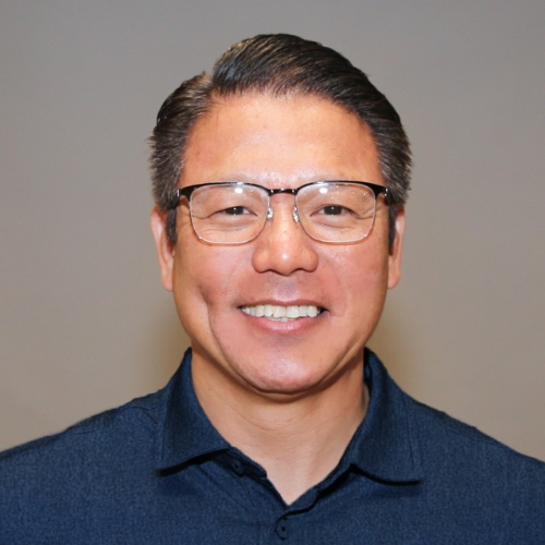 Picture of a man with glasses in a dark blue shirt with collar in front of a gray backgorund.
