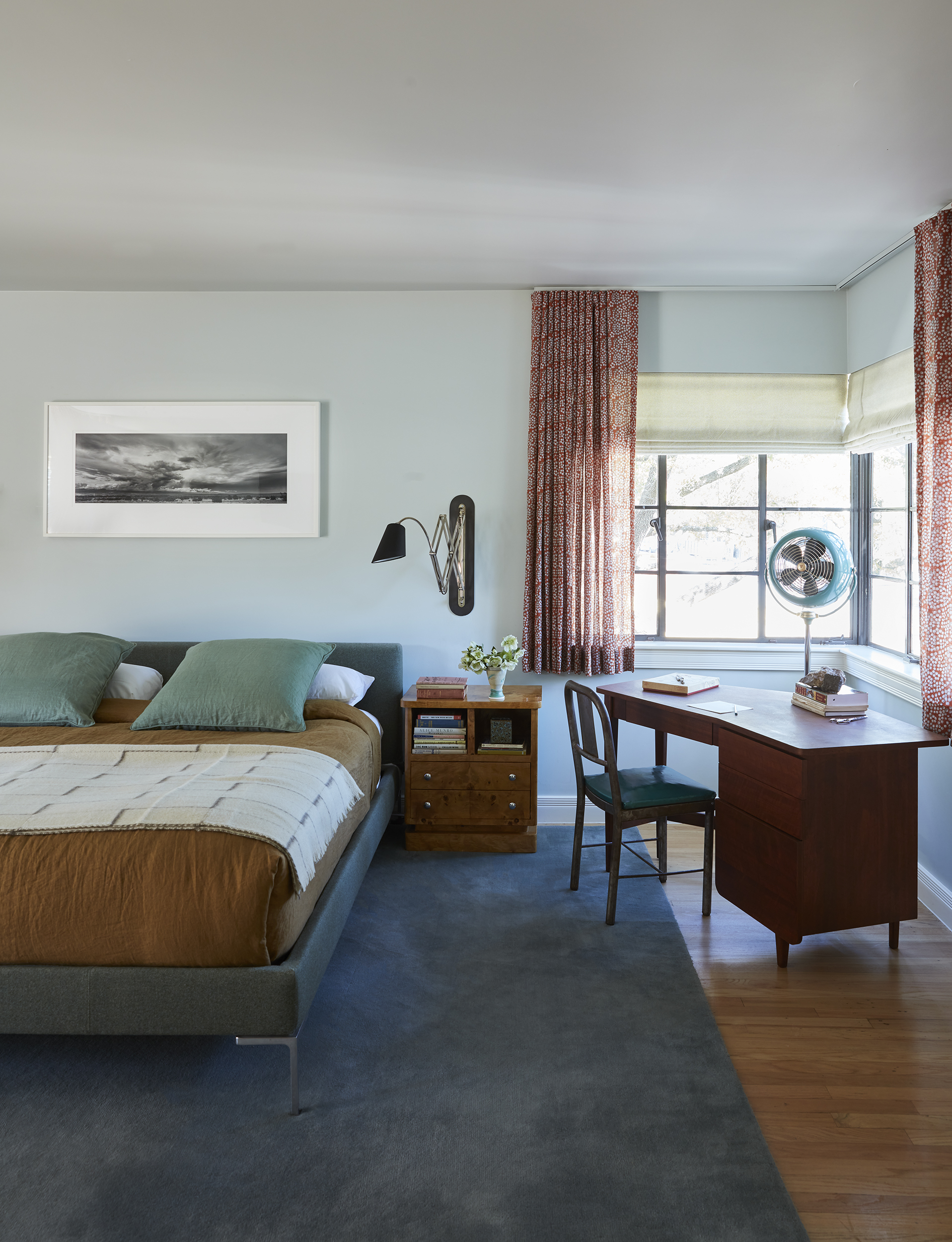 Image of a large, light blue bedroom with a bed in the middle and a wooden desk in the corner. The bed is blue with a brown duvet and blue -green pillow. There are windows above the desk with short, red curtains.