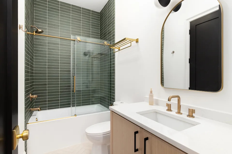 A bathroom with a wooden washing table, a golden hardware and a tap and a shower with green U -Bahn tiles.