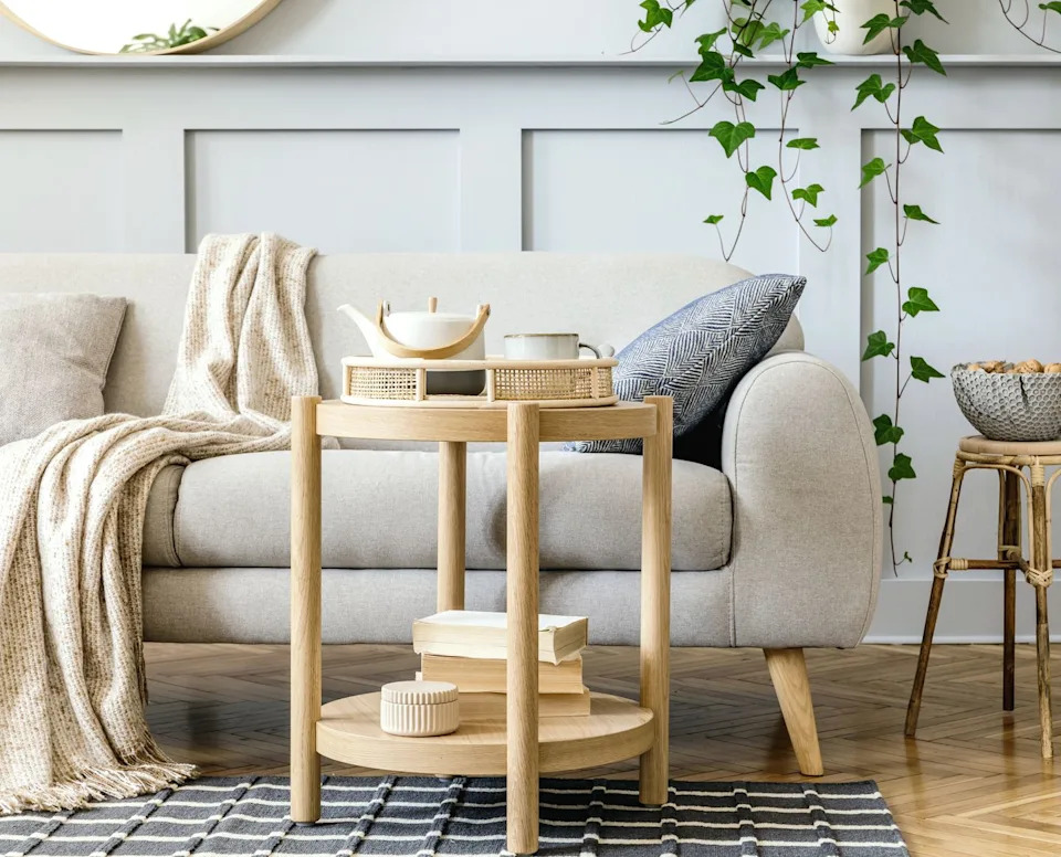 Scandinavian living room -interior with design gray sofa, wooden coffee table, tropical plants, shelf, mirror, furniture, checkered pillow, teapot, book and elegant personal accessories in home culture.
