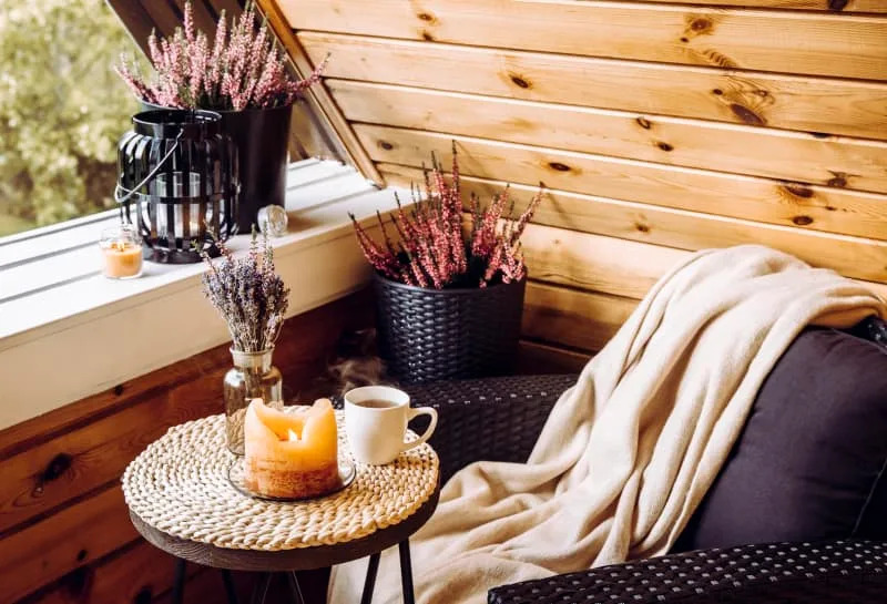 Nice autumn home arrangement. Tiny wooden cabin balcony with heather blum in the pot, lavender in a bottle vase, candlelight flame, soft beige plaid waiting for comfortable garden furniture chair