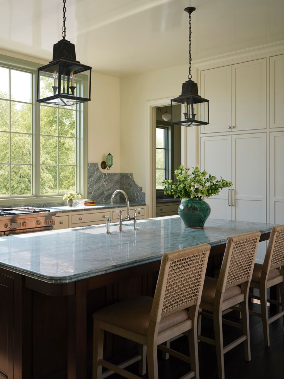 Elegant kitchen with marble Island, woven chairs and trailer lights over a picturesque window view.