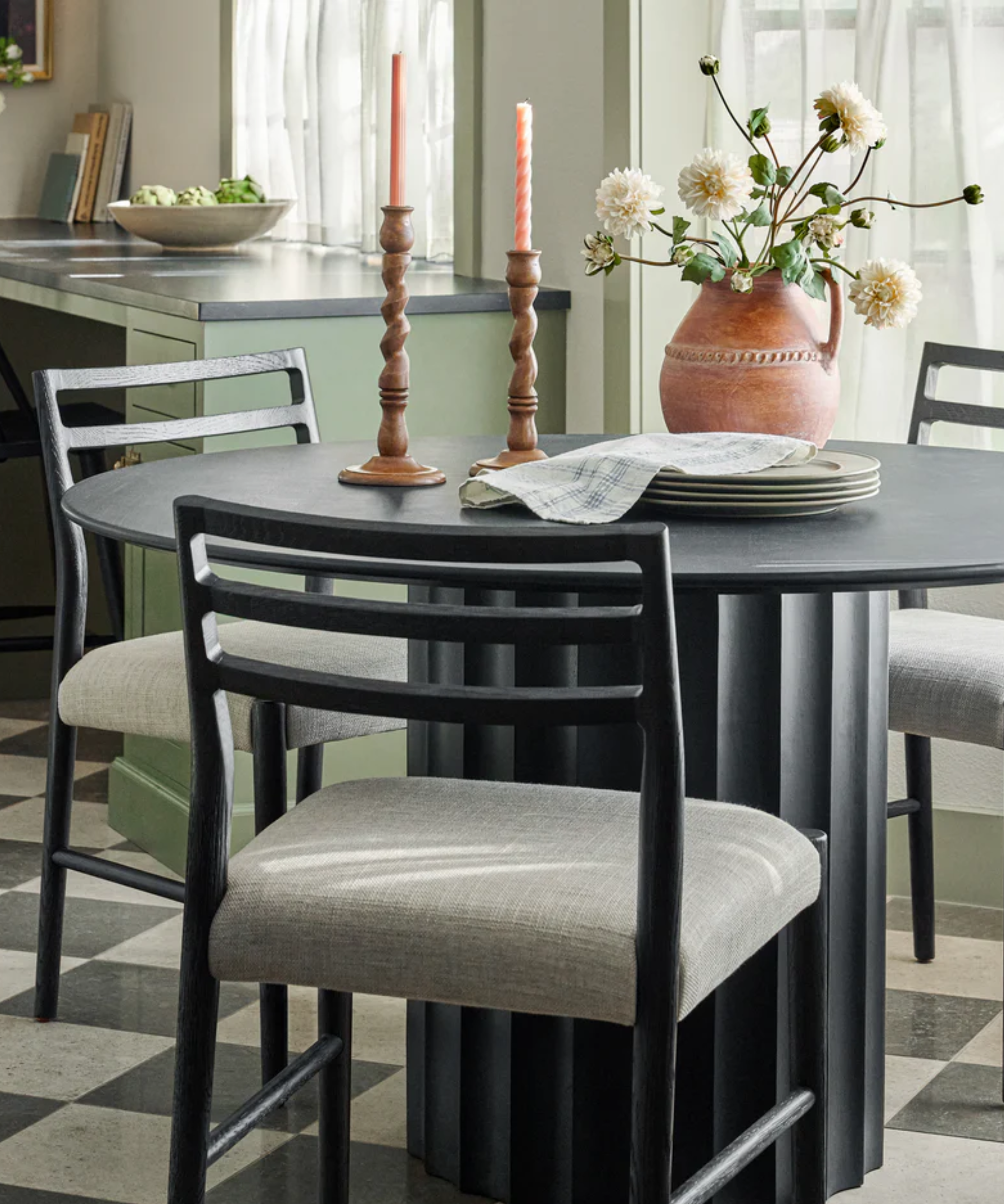 A dining room with a black dining table and eating chairs, twisted wooden candlesticks, a set of plates and a vase that holds flowers