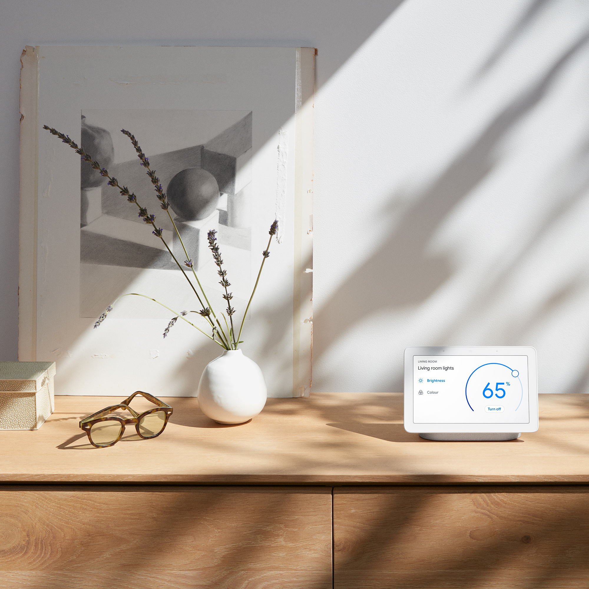 Google Home Hub on a wooden sideboard with different articles such as a vase with lavender springs and framed art print and glasses next to it