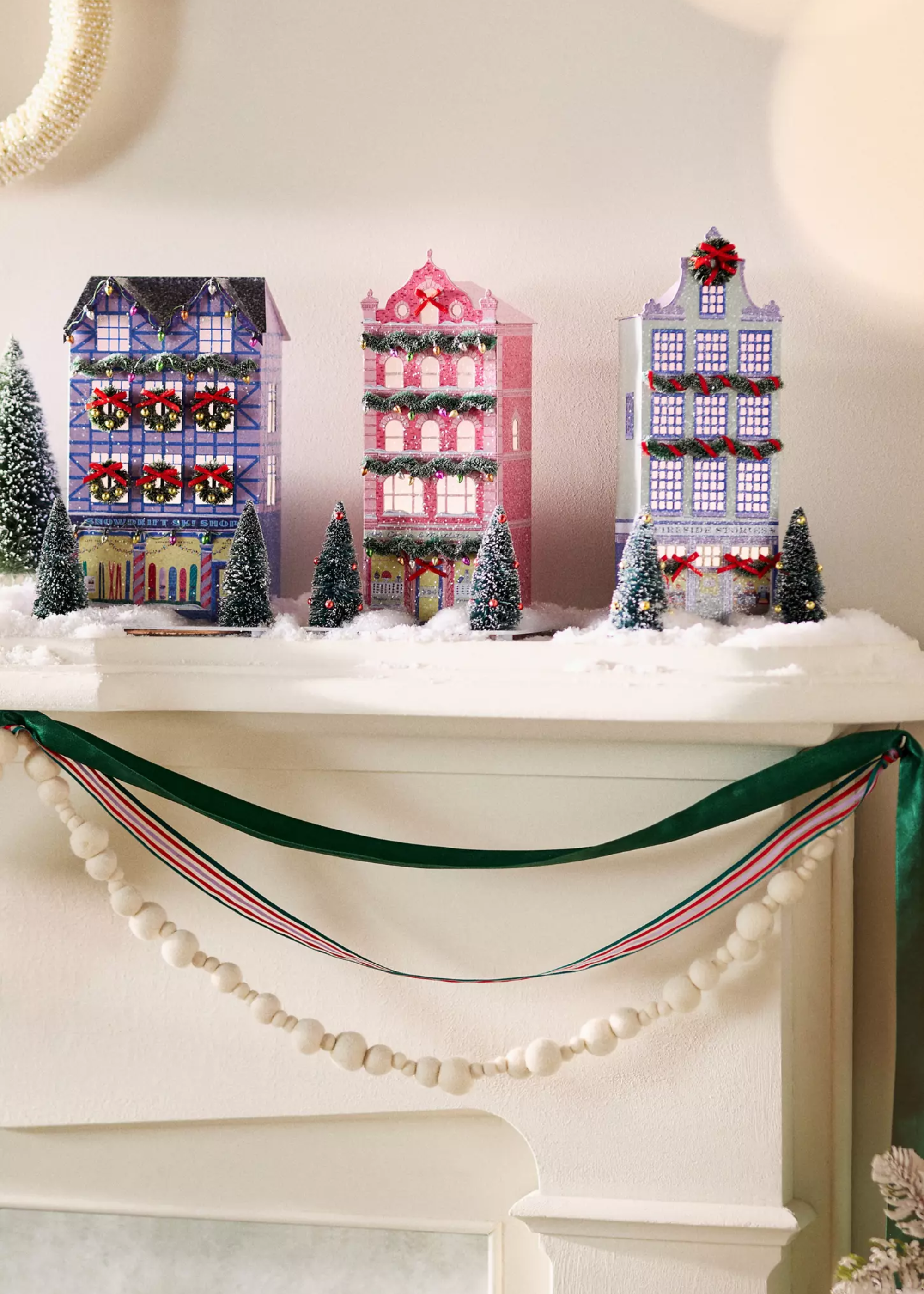 Anthropology Colored Christmas Village on a Kaminsims with band decorations underneath, false snow on the fireplace and miniature frost and decorated Christmas trees