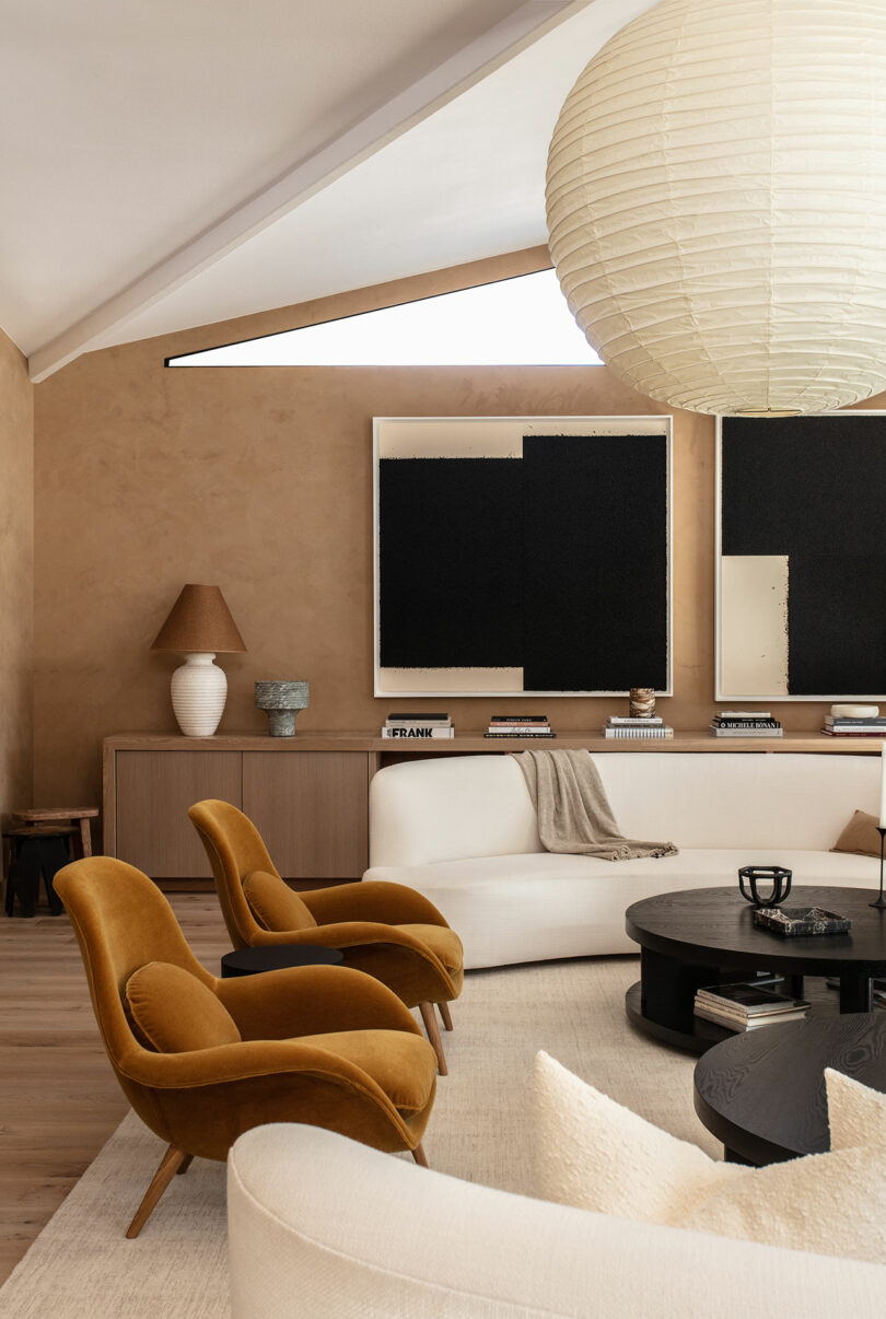 Modern living room with mustard velvet chairs, white curved sofas, round black coffee table, paper lantern trailer and neutral brown walls with abstract black and white art over a console.