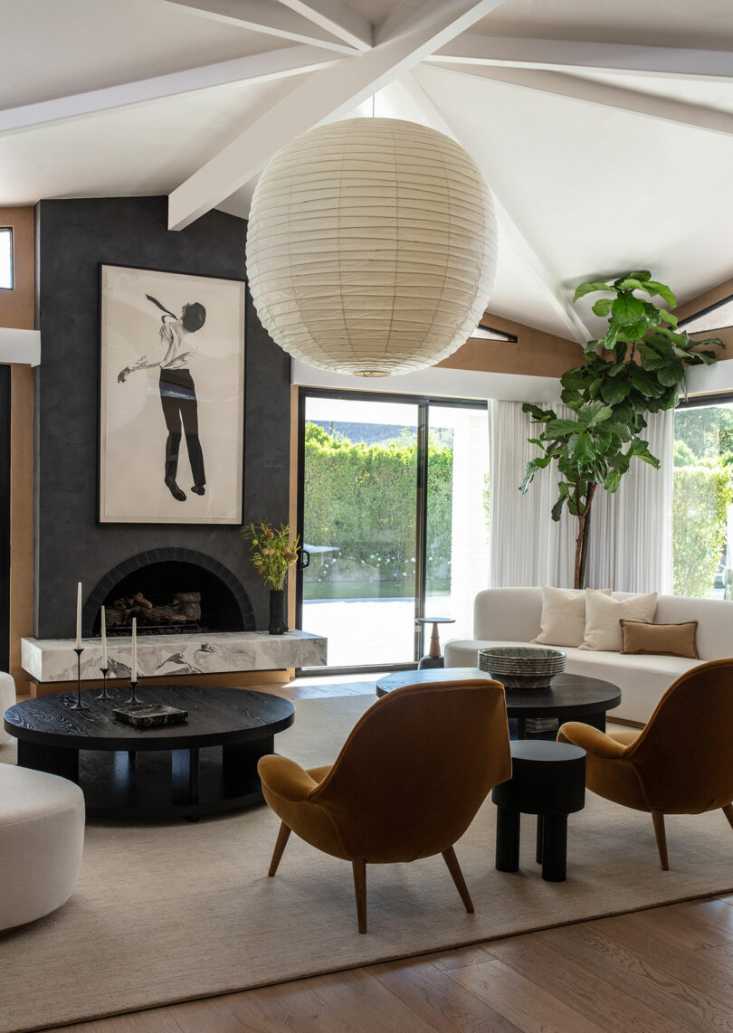 Modern living room with mustard yellow armchairs, a large paper lantern light, a black round coffee table, artwork over a fireplace and a high inner plant through bottom and cover windows.