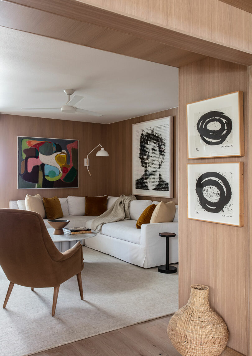 A modern living room with wooden walls, white clippings, brown chair, abstract and portrait graphics, small tables and a woven basket on the floor.