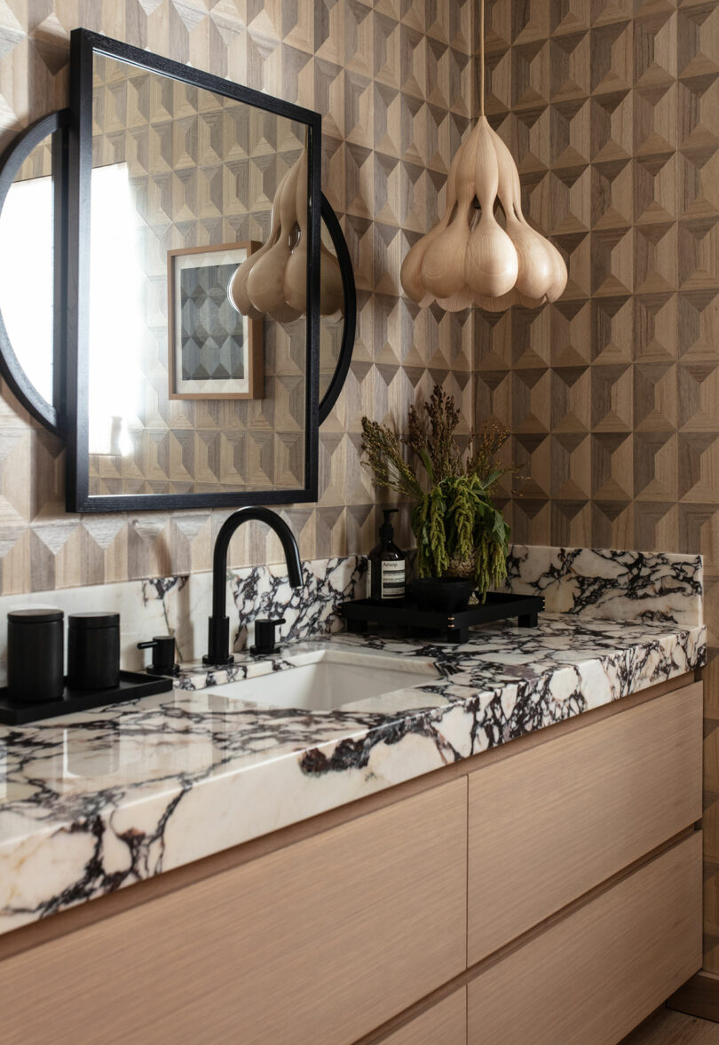 A modern bathroom with a worktop with a black and white marble, light wooden cabins, geometric wall tiles, a black frame mirror, a sculptural trailer and decorative plants.
