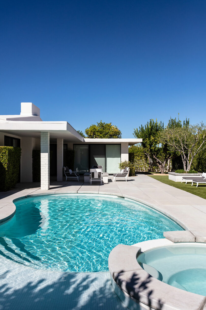 Modern house with a white outer and large windows with a round swimming pool and a spa under a clear blue sky with terrace furniture and surrounding greens.